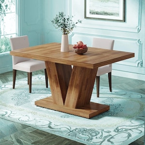 May include: A wooden dining table with a unique V-shaped base. The table is rectangular and has a light brown finish. Two white chairs are positioned on either side. A vase with greenery and a bowl of fruit are on the table.