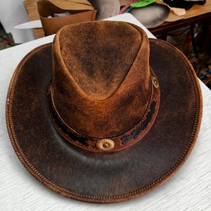 Könnte beinhalten: Ein Cowboyhut aus braunem Leder mit breiter Krempe und strukturierter Krone. Der Hut hat dekorative Metallakzente und ein Lederband. Ein klassisches Western-Accessoire, das vor Sonne schützt und Stil bietet.