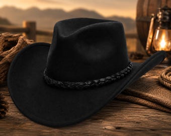 May include: A black cowboy hat with a braided band, resting on a wooden surface. The hat has a wide brim and a classic crown shape. The background features a rustic setting with a wooden fence and a lantern.