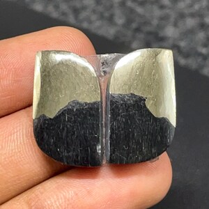 May include: A polished gemstone cabochon featuring a distinct split design. The upper portion displays a metallic, reflective gold-toned pyrite surface, while the lower section consists of a matte, textured black material with vertical striations.
