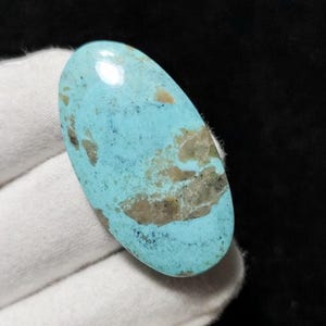 Puede incluir: Una piedra preciosa ovalada de turquesa con una superficie pulida. La piedra presenta un tono azul vibrante con patrones de matriz marrón y beige. La piedra preciosa se sostiene en una mano enguantada blanca sobre un fondo negro.