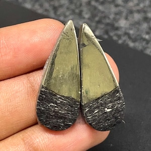 May include: Two teardrop-shaped cabochons featuring a dual-tone appearance. The upper portion of each piece is a smooth, metallic gold-toned pyrite, while the lower section consists of a textured, matte black material with visible horizontal striations.