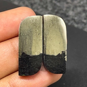 May include: Two matching rectangular stone cabochons with rounded top corners. Each piece features a distinct horizontal color split, with a metallic, brass-toned upper section and a textured, matte black lower section, creating a sharp contrast.