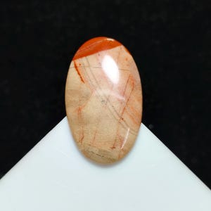 Puede incluir: Una gema ovalada con superficie pulida. La piedra presenta una mezcla de tonos beige, beis y rojo-anaranjado, con finas vetas rojas. La parte superior es de color rojo-anaranjado sólido.