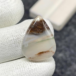 May include: A teardrop-shaped polished gemstone featuring a smooth, translucent surface with creamy white and grey tones. The stone displays distinct horizontal bands of earthy brown inclusions and a subtle, thin line of pale green running across the center.