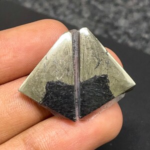 May include: A triangular-shaped gemstone featuring a metallic gold-colored pyrite surface with a distinct matte black mineral inclusion at the base. The stone is divided by a vertical line, creating a symmetrical appearance with sharp edges.
