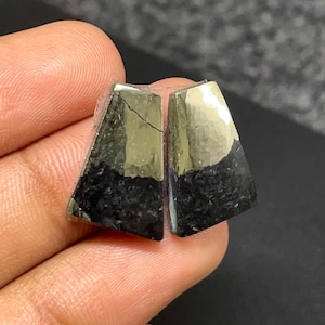 May include: Two trapezoidal stones featuring a dual-tone appearance with a metallic, reflective gold-colored upper section and a matte, textured black lower section. The stones have a distinct geometric shape with straight edges and a smooth surface.