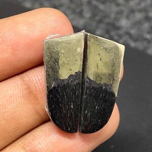May include: A polished shield-shaped gemstone featuring a dual-tone appearance. The upper section displays a metallic, reflective gold-toned pyrite surface, while the lower portion consists of a dark, textured black material with fine vertical striations.