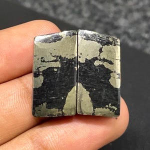May include: Two rectangular cabochons featuring a metallic gold pyrite pattern embedded within a solid black matrix. The stones have a smooth, polished surface with straight edges and a flat top, displaying a distinct contrast between the dark base and the reflective, irregular gold-toned mineral inclusions.