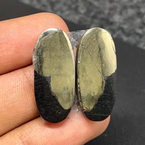May include: Two oval-shaped cabochons featuring a dual-tone appearance with a metallic gold-colored pyrite section and a contrasting matte black section. The stones have a smooth, polished surface with visible natural fissures and distinct color separation.