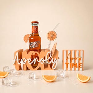 Könnte beinhalten: Eine Flasche Aperol Spritz steht in einem hellorangen Holzhalter mit dem Wort Aperoly in weißer Schreibschrift. Ein durchsichtiger Glasstrohhalm mit einem Orangenscheiben-Ausschnitt ist enthalten. Ein Schild trägt die Aufschrift Aperol TIME.