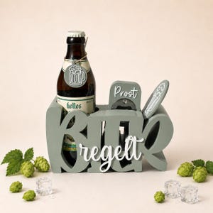 Könnte beinhalten: Ein salbeigrüner Flaschenhalter aus Holz mit dem weißen Schriftzug "Bier regelt" und "Prost" sowie einem integrierten Flaschenöffner. Eine braune Glasbierflasche mit "Helles"-Etikett und einem runden Anhänger mit Bierkrug-Motiv steht darin.