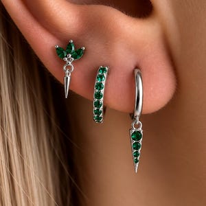 May include: Three silver-toned earrings featuring green stones. One earring has a three-leaf design with a dangling spike, one is a small hoop set with green stones, and the third is a hoop with a dangling, triangular, green-stone-set charm.