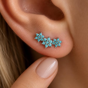 May include: A silver ear climber earring featuring three connected flower-shaped clusters set with small, round, bright turquoise stones. The earring follows the curve of the earlobe, with each flower composed of six petals surrounding a centre stone.