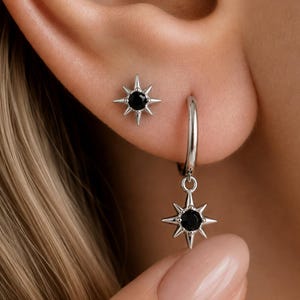 May include: A silver-toned stud earring featuring a black circular stone surrounded by eight pointed rays in a starburst shape, paired with a matching silver-toned hoop earring that has a dangling starburst charm with a central black stone.