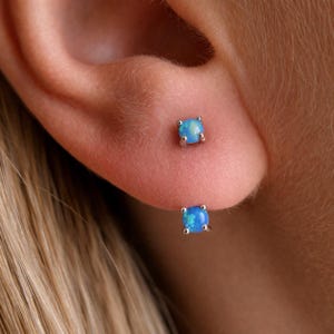 Puede incluir: Primer plano de dos pequeños pendientes de botón cuadrados. Cada pendiente presenta una piedra opalina azul claro engastada en metal plateado. Los pendientes están colocados en una oreja, centrándose en la joyería.