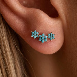 Può includere: Orecchini a bottone floreali color oro con pietre color turchese. Gli orecchini presentano tre gruppi di fiori, ciascuno con più pietre piccole e rotonde. Gli orecchini sono posizionati sulla pelle di un orecchio.