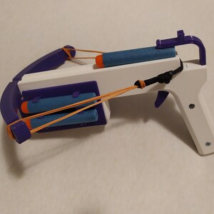 Pode incluir: Uma besta de brinquedo branca e roxa com elásticos laranja e dardos de espuma azuis. A besta tem um gatilho e uma pega. O arco é roxo e o corpo é branco.