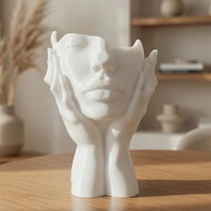 Könnte beinhalten: Eine weiße Vase in Form eines menschlichen Gesichts, bei der zwei Hände die Wangen umschließen. Die Skulptur hat eine glatte, matte Oberfläche mit ausgeprägten Gesichtszügen wie geschlossenen Augen, Nase und Lippen sowie einer Öffnung.