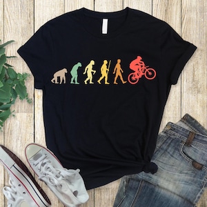 Può includere: T-shirt nero con una grafica evolutiva colorata, che mostra la progressione dalla scimmia al biker. Il design utilizza una sfumatura di colori, dal giallo al rosso. La maglietta è abbinata a jeans blu e scarpe da ginnastica bianche.