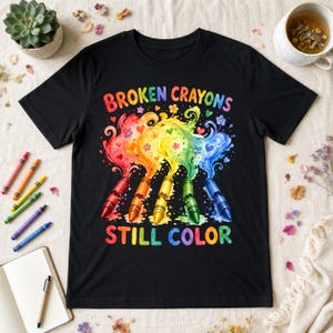 Könnte beinhalten: Ein schwarzes Kurzarm-T-Shirt mit einer Grafik aus fünf zerbrochenen Buntstiften in Regenbogenfarben, wirbelnden Farbspritzern, kleinen Herzen und Blumen. Der Text "BROKEN CRAYONS" steht über der Grafik und "STILL COLOR" darunter in bunten Buchstaben.