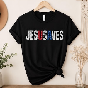 Könnte beinhalten: Schwarzes T-Shirt mit dem Text "JESUS SAVES" in abgenutzten weißen Buchstaben. Die Buchstaben "U" und "S" sind rot mit weißen Sternen, und der Buchstabe "A" ist blau mit weißen Sternen. Das T-Shirt hängt an einem Holzbügel.