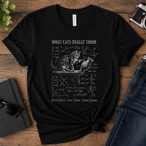 Könnte beinhalten: Schwarzes T-Shirt mit einem Aufdruck mit der Aufschrift "WHAT CATS REALLY THINK". Die Grafik zeigt eine Katze, eine Vase und mathematische Gleichungen. Die Namen Galileo Galilei, Isaac Newton und Robert Goddard sind ebenfalls auf dem T-Shirt abgedruckt.