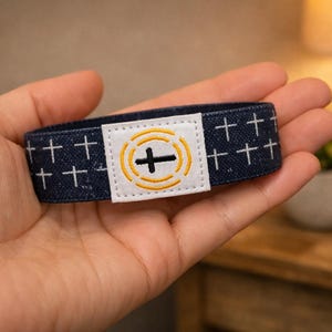 May include: A dark blue elastic fabric wristband featuring a repeating white cross pattern. A rectangular white patch is centered on the band, displaying an embroidered black cross surrounded by two concentric yellow circular lines.