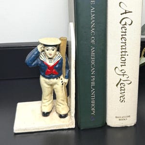 May include: A decorative sailor bookend in blue, red, and white holds two books. The bookend features a sailor figurine saluting. The books are titled "The Almanac of American Philanthropy" and "A Generation of Leaves."