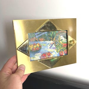 May include: A gold-colored metal picture frame with a diamond-shaped design. The frame holds a colorful painting of a still life with fruit and vegetables. The frame is being held by a hand.