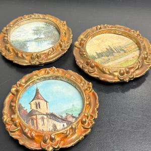 May include: Three circular wall frames with ornate, distressed gold-tone edges. Each frame contains a small, detailed landscape print: one shows a river scene, another a field with trees, and the third depicts a stone church building under a blue sky.