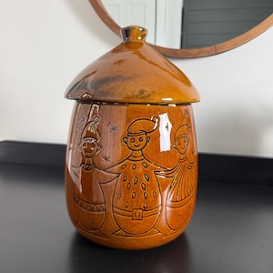 May include: A glossy, amber-colored ceramic canister with a matching lid. The exterior features a dark brown etched illustration of three figures wearing hats and patterned clothing, standing side-by-side with small star accents surrounding them.