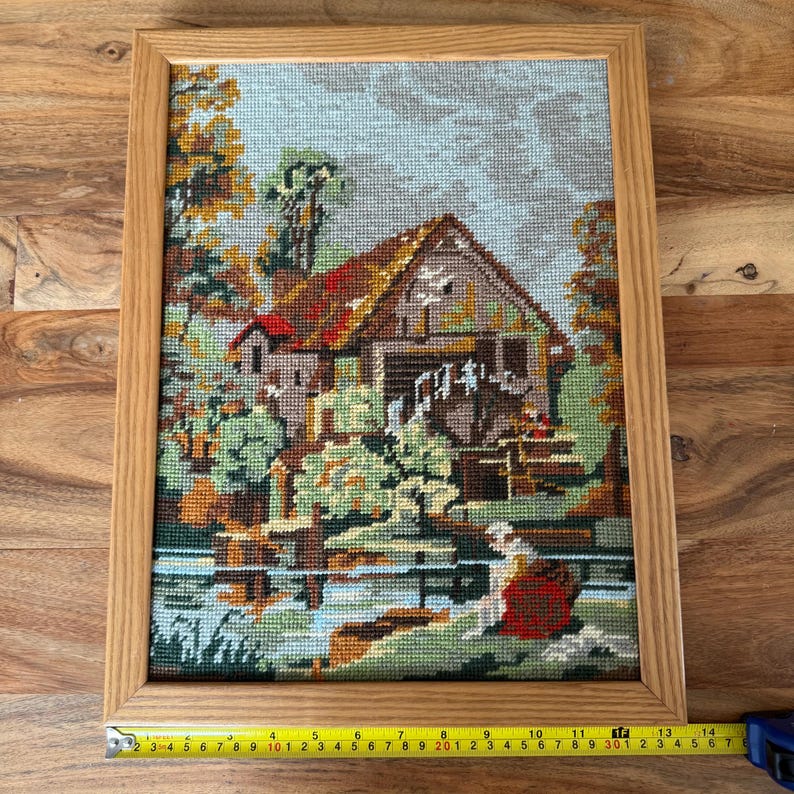 May include: Framed needlepoint art depicting a rural scene with a watermill, trees, and a body of water. The artwork features a wooden frame and a colorful, detailed design. The scene includes a building with a red roof and a water wheel.