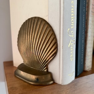 May include: A brass bookend shaped like a scallop shell with prominent vertical ridges and a smooth, curved base. The metallic object is positioned next to a white book spine with gold lettering that reads "erution of Leaves".