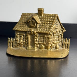 May include: A brass doorstop shaped like a small cottage with a textured roof, a central chimney, and a picket fence. The house features detailed stone walls, a front door, and windows with visible panes, all cast in a metallic gold finish.