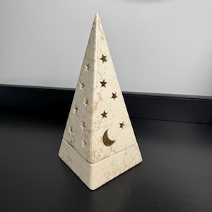 May include: A triangular, pyramid-shaped decorative object with a cream-colored base featuring a marbled, veined texture. The surface is perforated with multiple cut-out star shapes of varying sizes and a single crescent moon shape near the base.