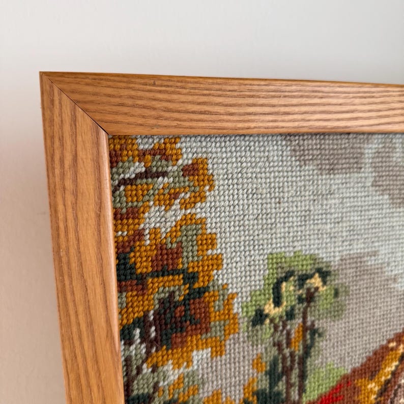 May include: A framed needlepoint artwork featuring a landscape scene with trees in shades of yellow, orange, and green. The artwork is encased in a light brown wooden frame, adding a classic touch to the piece.