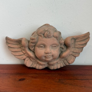 May include: A terracotta cherub head sculpture with detailed wings. The cherub has a serene expression and curly hair. The sculpture is a light brown color and is likely made of clay or a similar material. The wings are spread out on either side of the head.