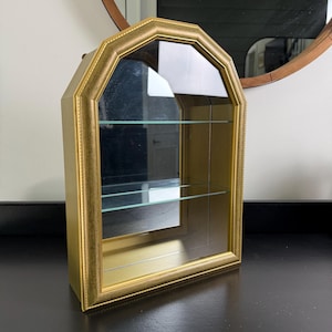 May include: A wall-mounted display cabinet with a gold-toned frame and an arched top. The cabinet features a mirrored back panel, a clear glass front door, and two interior glass shelves for displaying small items within the gold-finished structure.