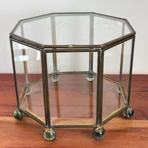 May include: An octagonal glass and brass display box. The clear glass panels are framed by brass-colored metal. The box has small, round, green glass feet. The top is also octagonal. The box is sitting on a wooden surface.