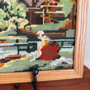 May include: Framed needlepoint art depicting a serene landscape scene. The artwork features a figure in a red garment near a body of water, with a bridge and buildings in the background. The frame is made of light-colored wood.
