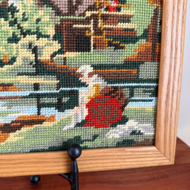 May include: Framed needlepoint art depicting a serene landscape scene. The artwork features a figure in a red garment near a body of water, with a bridge and buildings in the background. The frame is made of light-colored wood.