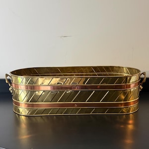 May include: A brass oval planter with copper bands and diagonal etched lines. The planter has rope handles on each end. The planter is a decorative item for indoor or outdoor use.
