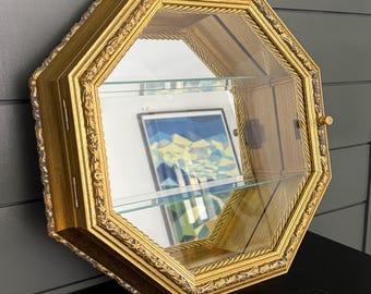 May include: An octagonal wall-mounted display cabinet featuring a gold-toned frame with ornate detailing, a hinged glass door with a small knob, and two interior glass shelves. The back of the cabinet is mirrored, reflecting the surrounding environment.