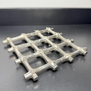 May include: A silver-colored metal trivet with a grid pattern. The trivet is made of interconnected, rounded bars, forming a square shape. The design is simple and elegant, suitable for placing hot dishes.