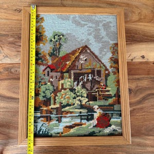 May include: A framed needlepoint tapestry depicting a rural scene with a watermill, trees, and a figure by the water. The tapestry is in a wooden frame. The image is approximately 17 inches tall.