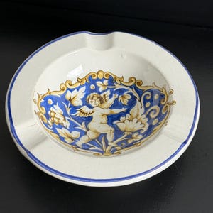 May include: A round, white ceramic ashtray with a blue and gold decorative design. The design features a cherub and floral elements. The rim of the ashtray is outlined in blue.