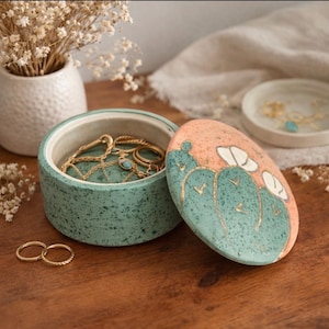 May include: A round, speckled, teal jewelry box with a matching lid. The lid is decorated with a cactus design in teal and gold against a peach background. Inside the box are gold necklaces and rings. Two gold rings sit on the wooden surface.