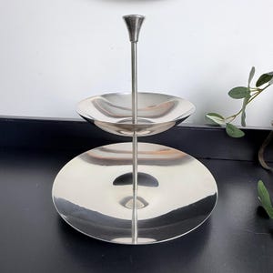 May include: A two-tiered, silver-toned serving stand. The stand features two circular trays connected by a central rod. The top tray is smaller than the bottom tray. The stand has a simple, elegant design, suitable for displaying desserts or appetizers.