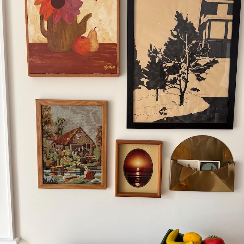 May include: A collection of framed art pieces and a brass wall pocket. The art includes a painting of a floral still life, a black and white landscape, a tapestry of a watermill, and a sunset scene. The brass pocket holds cards.